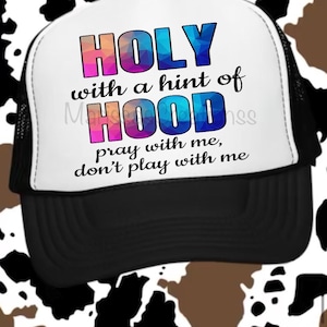 May include: A black and white trucker hat with the text "HOLY with a hint of HOOD pray with me, don't play with me" in colorful lettering. The hat has a white front panel and a black mesh back, with a curved brim.