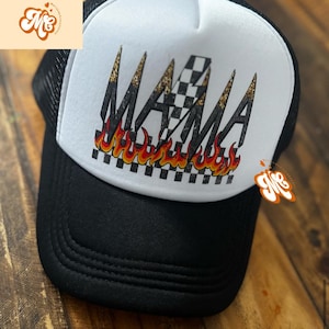 May include: Black and white trucker hat with the word "MAMA" in a bold, stylized font. The design includes a checkered pattern, flames, and gold accents. The hat has a mesh back and a curved brim.