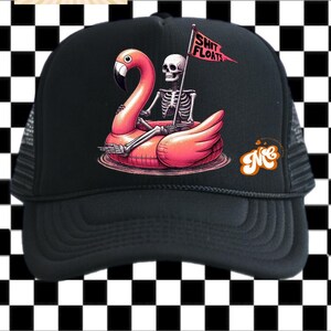 May include: Black trucker hat featuring a pink flamingo pool float with a skeleton rider holding a flag that reads "SHIT FLOATS." The hat has a black mesh back and a curved brim. The design is set against a black and white checkered background.