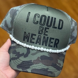 May include: A camouflage trucker hat with the text "I COULD BE MEANER" in black. The hat features a white pearl bead trim along the brim and a mesh back.