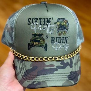 May include: An olive green trucker hat with a camouflage brim and mesh back. The front panel features a graphic of an off-road vehicle and a stylized head with a bow, accompanied by the black text "SITTIN' Pretty & RIDIN' Dirty". A gold chain trim sits across the base of the front panel.