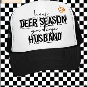 May include: A black and white trucker hat with the text "hello DEER SEASON goodbye HUSBAND" in black. The hat has a white front panel and a black brim and mesh back. A small orange butterfly graphic is on the right side of the hat.
