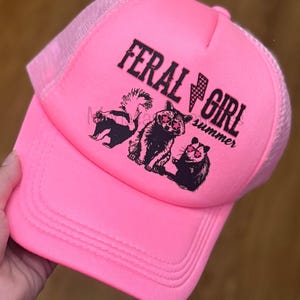 May include: Bright pink trucker hat with a mesh back. The front of the hat features the text "FERAL GIRL" with a lightning bolt graphic and the word "summer". Below the text are illustrations of a skunk, raccoon, and a rodent, all wearing sunglasses.