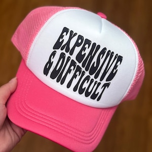 May include: A pink trucker hat with a white front panel. The hat features the text "EXPENSIVE & DIFFICULT" in black, stylized lettering. The hat has a pink brim and mesh back.
