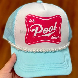 May include: A light blue and white trucker hat featuring a mesh back and a solid front panel. The front displays a pink graphic with the text 'it's Pool time' in a stylized font. A row of white faux pearls lines the base of the front panel.