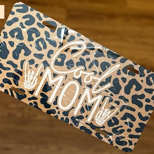 May include: License plate with a tan and black leopard print background. The words "Cool MOM" are in white, stylized lettering. Skeleton hands are used as the "O" in "Cool" and the "O" in "MOM".