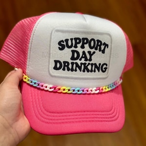 May include: A bright pink trucker hat featuring a white front panel with the black text SUPPORT DAY DRINKING printed in a bold font. The hat includes a vibrant, multi-colored plastic chain trim across the base of the front panel and mesh sides.