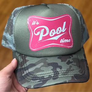May include: An olive green trucker hat featuring a camouflage pattern on the brim and mesh back panels. The front panel displays a pink, rectangular graphic with rounded corners containing the white text it's Pool time in a stylized font.