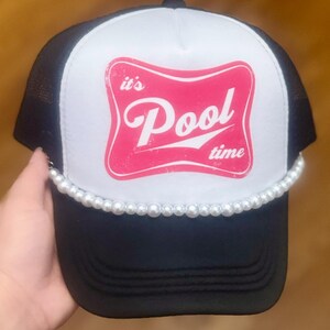 May include: A black and white trucker hat featuring a pink graphic label on the front panel with the white text "it's Pool time". The hat is accented with a single strand of white faux pearls lining the base of the front crown above the brim.