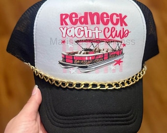 May include: A black and white trucker hat featuring a gold chain across the base of the front panel. The white front panel displays a pink graphic of a pontoon boat with the text Redneck Yacht Club in pink lettering and small pink stars.