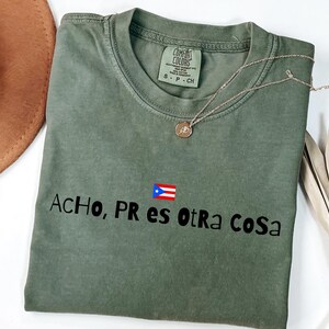 Può includere: T-shirt verde oliva con la scritta "ACHO, PR es otra cosa" e una grafica della bandiera portoricana. Una collana d'oro con un ciondolo è appoggiata sulla maglietta. La maglietta è piegata e esposta su una superficie bianca.