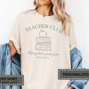 Puede incluir: Camiseta de color marfil con un gráfico verde claro. El gráfico incluye las palabras "TEACHER CLUB", una manzana y una pila de libros. Debajo de los libros, se lee "Helping little minds grow" y "MRS. SMITH".