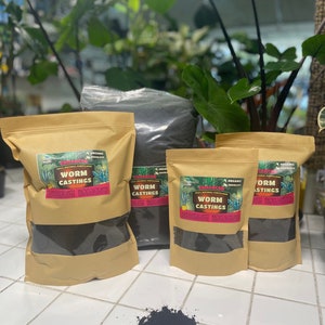 May include: Four brown paper bags filled with dark brown worm castings. The bags have a label that says "Enhancer 100% Natural Fertilizer Worm Castings". There is a small pile of dark brown worm castings on a white tiled surface.