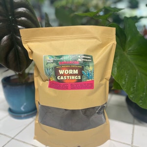 May include: A tan bag of "Worm Castings" organic fertilizer. The bag has a clear window showing the dark, granular contents. The label includes the words "Enhancer" and "Odorless". The background features green plants.