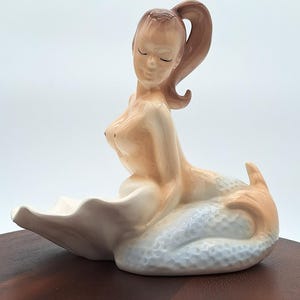 May include: A ceramic mermaid figurine with a light skin tone, brown ponytail, and closed eyes. The mermaid is seated in a white seashell with a textured, light blue tail. The figurine is on a dark brown surface.