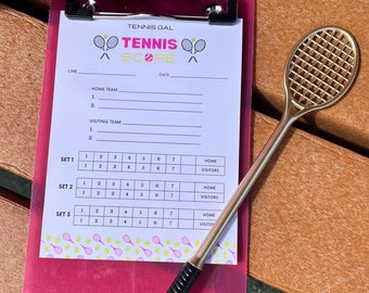 May include: A pink clipboard with a white printable tennis scorecard. The scorecard has the title "Tennis Gal Tennis Score" and includes spaces to record the home and visiting team names, line numbers, date, and scores for three sets. A gold-coloured miniature tennis racket is lying on top of the clipboard.