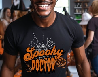 May include: A person wearing a black witch hat and a black t-shirt with the text "Spooky Doctor" in orange and white. The shirt also features a white spiderweb design and a small orange first aid kit graphic.