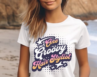 May include: White t-shirt with a pink and purple graphic that says "One Groovy Hair Stylist" with a star and polka dots.