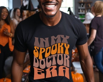 May include: A man wearing a black witch hat and a black t-shirt with the text "In My Spooky Doctor Era" in orange and brown letters. He is smiling and surrounded by other people at a Halloween party.