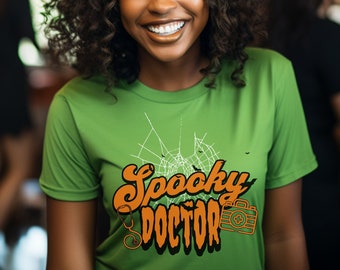 May include: A woman wearing a green t-shirt with a spiderweb design and the text "Spooky Doctor" in orange. She is wearing black cat ears and smiling.