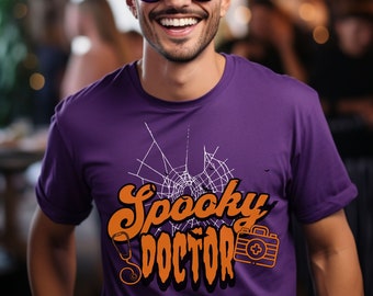 May include: A purple t-shirt featuring a white spiderweb design and the text "Spooky Doctor" in orange. The shirt also includes a small orange medical bag with a white cross.