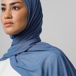 May include: A blue hijab, covering the head and shoulders. The fabric has a textured, ribbed appearance. The head covering is a solid, medium blue colour. The background is a plain, light grey.