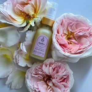 May include: A bottle of facial elixir with a gold cap and a white pump, surrounded by pink and white roses. The label reads "noita botanicals rose flower dew facial elixir". The roses have soft pink and yellow hues.