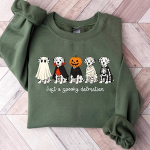 May include: An olive green crewneck sweatshirt featuring a graphic of five Dalmatian dogs in various Halloween costumes, including a ghost, vampire, pumpkin head, mummy, and skeleton. The text below the dogs reads, Just a spooky dalmatian.