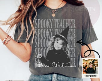 May include: A charcoal gray short-sleeve t-shirt featuring a repeating distressed white text graphic that reads SPOOKY TEACHER. The center graphic shows a black and white photo of a child in a witch hat with the white cursive text Mrs. Wilson.