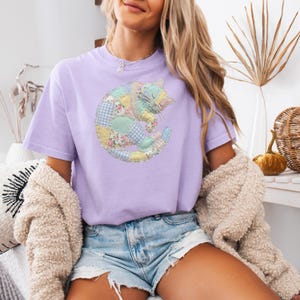 May include: A light purple t-shirt featuring a patchwork cat design in pastel colors. The cat is curled up, with a floral and gingham pattern. The shirt is short-sleeved and has a relaxed fit, perfect for casual wear.