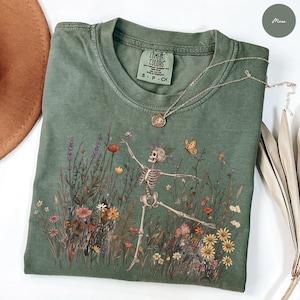 Puede incluir: Una camiseta de cuello redondo color verde musgo con un estampado gráfico de un esqueleto bailando en un campo de flores silvestres coloridas y una mariposa. La prenda tiene una etiqueta Comfort Colors en el cuello y un collar dorado encima.