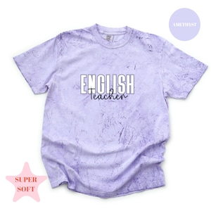 May include: A light purple t-shirt with a marbled effect. The shirt features the words "ENGLISH Teacher" in white block letters with a cursive "Teacher" underneath. A pink star with "SUPER SOFT" is in the lower left corner, and a purple circle with "AMETHYST" is in the upper right.
