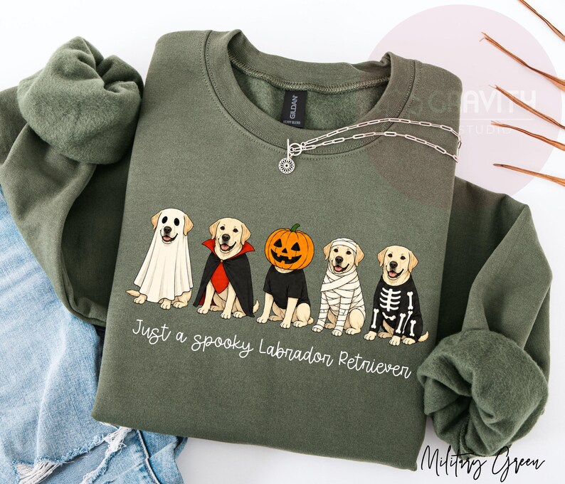Może przedstawiać: Oliwkowa bluza z okrągłym dekoltem z grafiką przedstawiającą pięć labrador&oacute;w w kostiumach na Halloween: ducha, wampira, dynię, mumię i szkielet. Pod psami znajduje się napis: Just a spooky Labrador Retriever.