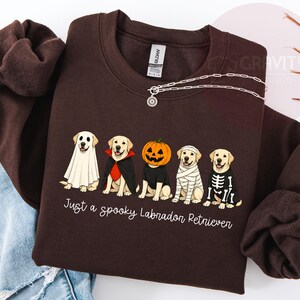 Może przedstawiać: Ciemnobrązowa bluza z okrągłym dekoltem z grafiką przedstawiającą pięć labrador&oacute;w w kostiumach na Halloween, w tym ducha, wampira, dynię, mumię i szkielet. Poniżej znajduje się napis, Just a spooky Labrador Retriever.