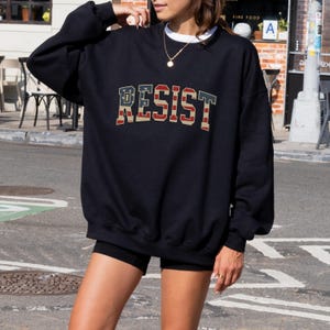 Puede incluir: Una sudadera azul marino con la palabra "RESIST" en un diseño rojo, blanco y azul en el pecho. La sudadera tiene mangas largas y cuello redondo. El texto está en negrita, estilo bloque.