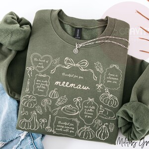May include: An olive green crewneck sweatshirt featuring a white line-art graphic with the text "meemaw," "thankful for you," and various phrases like "the heart of our family" surrounded by illustrations of pumpkins, bows, and food.