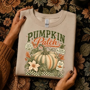 May include: A beige crewneck sweatshirt featuring a graphic print with the text "PUMPKIN Patch" in green and orange. The design includes a large pumpkin, leaves, a flower, and small labels reading "FRESH FROM THE PATCH," "FARM FRESH," and "LOCALLY GROWN."