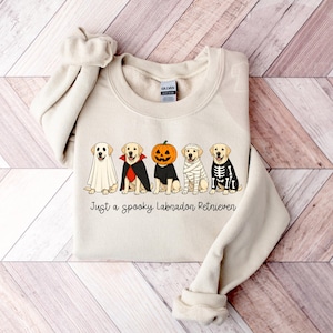 Może przedstawiać: Kremowa bluza z okrągłym dekoltem ozdobiona grafiką przedstawiającą pięć labrador&oacute;w w r&oacute;żnych kostiumach na Halloween, w tym ducha, wampira, dynię, mumię i szkielet. Tekst pod psami brzmi, Just a spooky Labradon Retriever.
