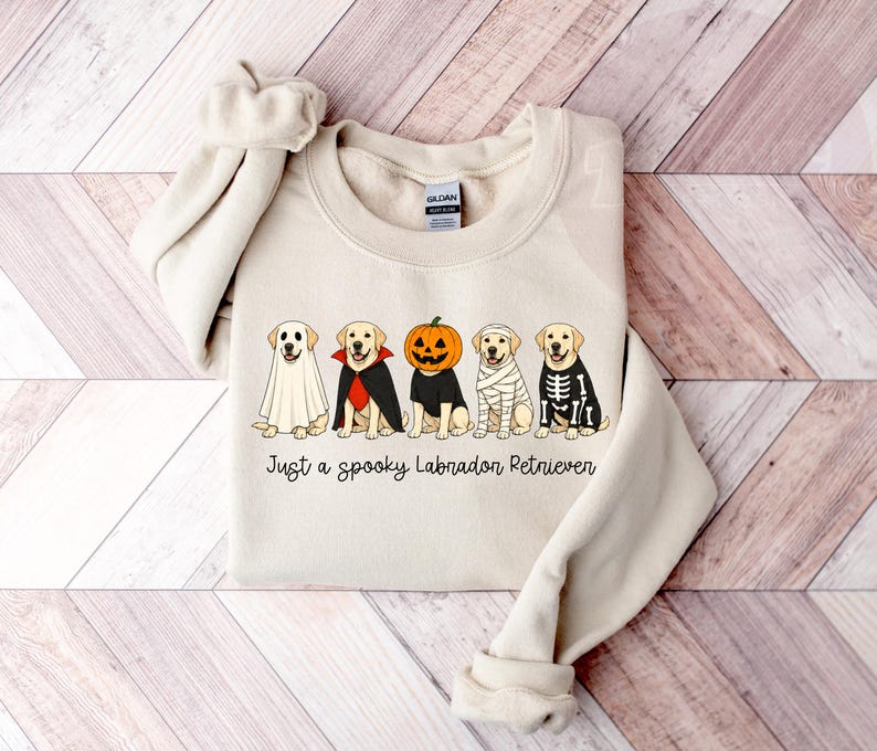 Może przedstawiać: Kremowa bluza z okrągłym dekoltem ozdobiona grafiką przedstawiającą pięć labrador&oacute;w w r&oacute;żnych kostiumach na Halloween, w tym ducha, wampira, dynię, mumię i szkielet. Tekst pod psami brzmi, Just a spooky Labradon Retriever.