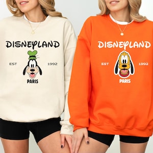 Puede incluir: Dos sudaderas, una crema y otra naranja, con el logo de Disneyland y la palabra "Par&iacute;s". La sudadera crema tiene un dise&ntilde;o de Goofy, y la naranja, Pluto. Ambas incluyen el texto "EST 1992."