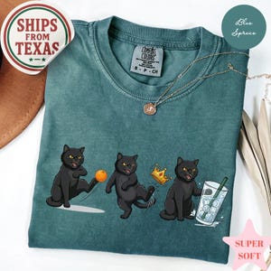 Puede incluir: Camiseta Comfort Colors de color verde azulado con un gr&aacute;fico de tres gatos negros. Uno patea una pelota, otro lleva una corona y el tercero sostiene una bebida. La camiseta tiene el texto "SHIPS FROM TEXAS".