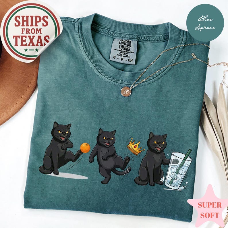 Puede incluir: Camiseta Comfort Colors de color verde azulado con un gr&aacute;fico de tres gatos negros. Uno patea una pelota, otro lleva una corona y el tercero sostiene una bebida. La camiseta tiene el texto "SHIPS FROM TEXAS".