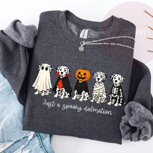 May include: A dark gray crewneck sweatshirt featuring a graphic of five Dalmatian dogs in Halloween costumes, including a ghost, vampire, pumpkin head, mummy, and skeleton. The text below the dogs reads, Just a spooky dalmatian.