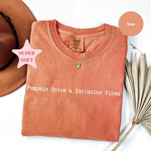 Może przedstawiać: Pomarańczowa bluza z okrągłym dekoltem z białym napisem "Pumpkin Spice & Inclusion Vibes". Złoty naszyjnik z wisiorkiem spoczywa na koszuli. Różowa grafika w kształcie gwiazdy głosi "Super Soft". Brązowy kapelusz i suszony liść palmy znajdują się w tle.
