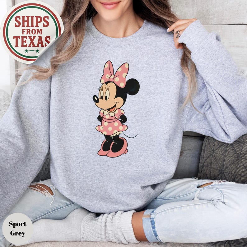 May include: Sport grey sweatshirt featuring a Minnie Mouse graphic. The cartoon character is wearing a pink polka dot dress and bow. The sweatshirt has a crew neck and long sleeves. The image also includes a "Ships From Texas" logo.