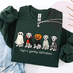 May include: A dark green long-sleeve sweatshirt featuring a graphic of five Dalmatian dogs in Halloween costumes, including a ghost, vampire, pumpkin head, mummy, and skeleton. The text below the dogs reads, Just a spooky dalmatian.