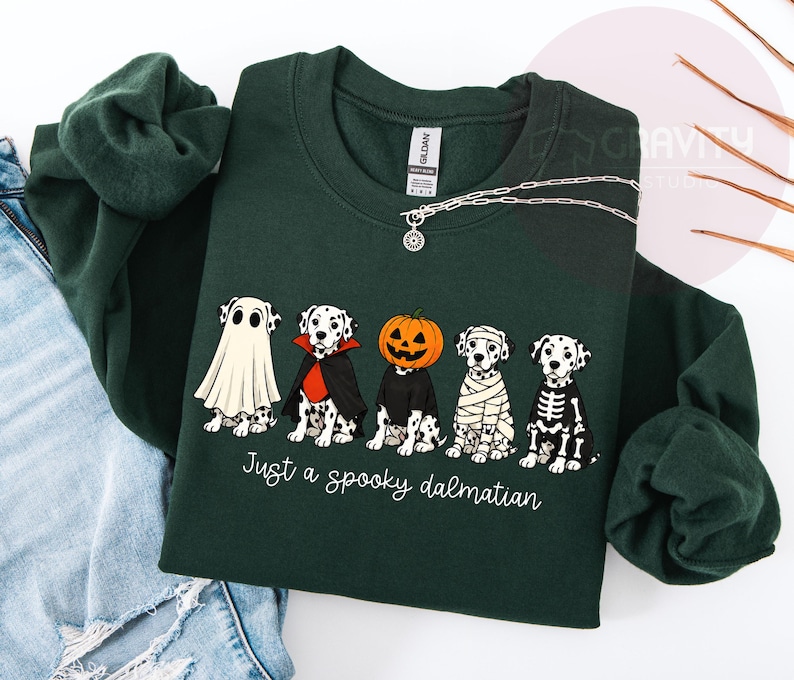 May include: A dark green long-sleeve sweatshirt featuring a graphic of five Dalmatian dogs in Halloween costumes, including a ghost, vampire, pumpkin head, mummy, and skeleton. The text below the dogs reads, Just a spooky dalmatian.