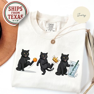 Puede incluir: Camiseta color marfil con tres ilustraciones de gatos negros. Los gatos se representan en varias poses juguetonas con una pelota, una corona y una bebida. La camiseta tiene una etiqueta Comfort Colors y una pegatina Ships From Texas.