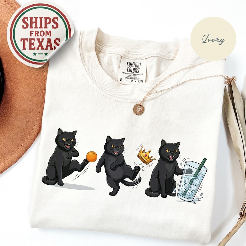 Puede incluir: Camiseta color marfil con tres ilustraciones de gatos negros. Los gatos se representan en varias poses juguetonas con una pelota, una corona y una bebida. La camiseta tiene una etiqueta Comfort Colors y una pegatina Ships From Texas.