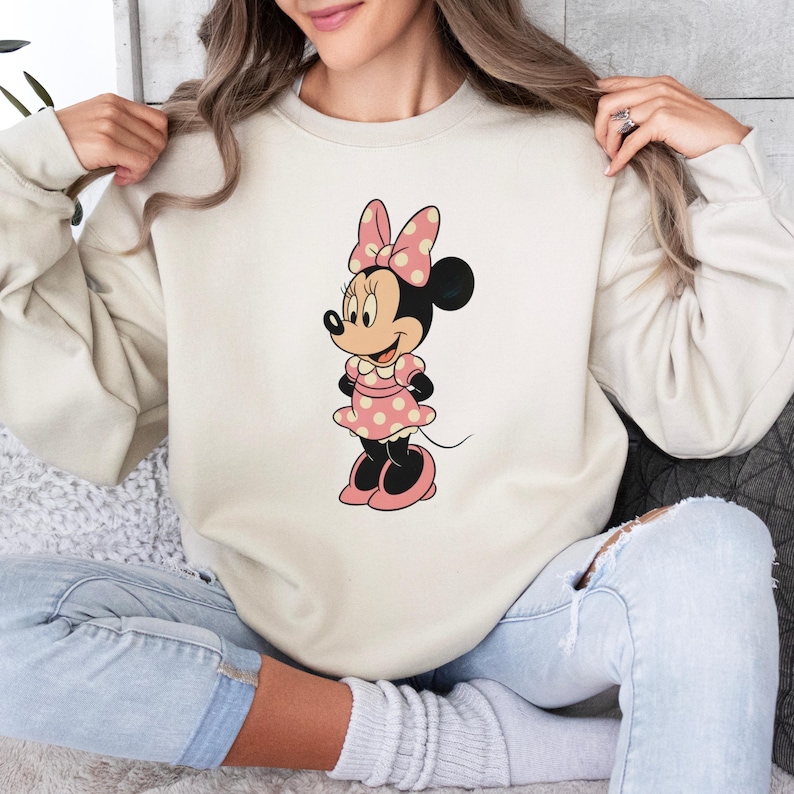 May include: Cream-colored sweatshirt featuring a Minnie Mouse graphic. The graphic shows Minnie Mouse in a pink polka-dot dress, bow, and shoes. The sweatshirt is a long-sleeved, crew neck style.